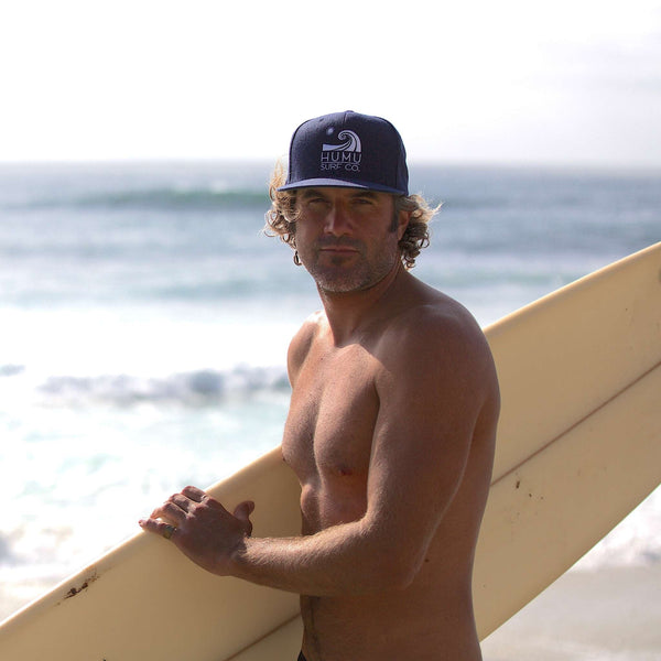 Surfer holding a longboard at the beach while wearing the navy Humu Wave Snapback Cap