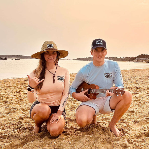 Man and woman wearing Humu Woody Bamboo Long-Sleeve Sun Shirts with a Classic Lifeguard Hat and Original Black Trucker Hat