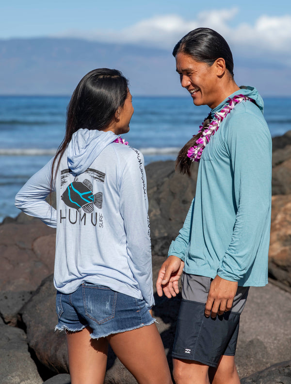 Two people wearing white and marine blue Humu Woody bamboo lightweight hoodies on a rocky shoreline by the ocean.