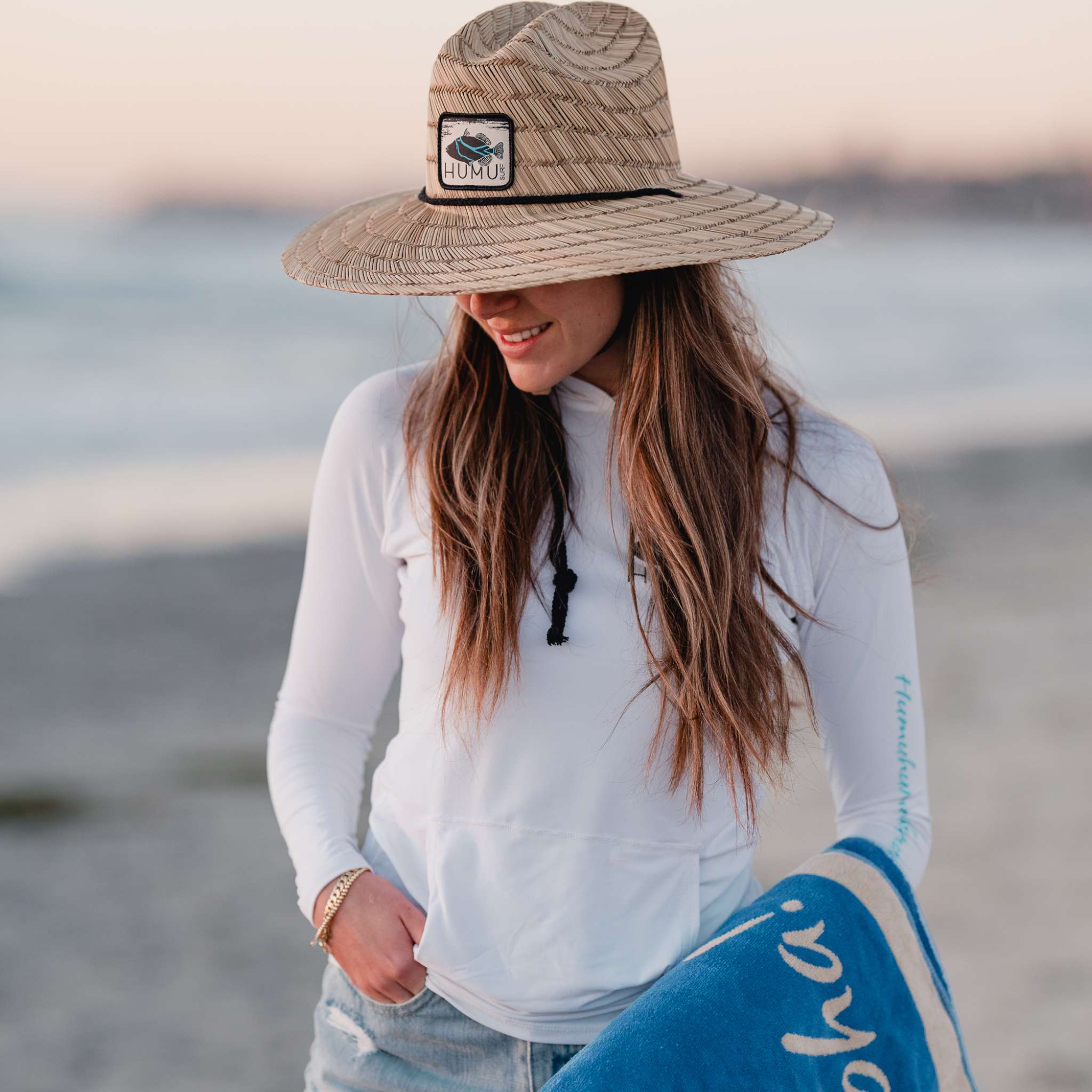 Woman wearing the Humu Classic Lifeguard Hat at the beach while carrying a blue Live With Aloha brand Humu Surf towel