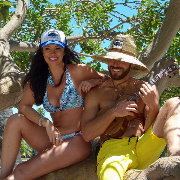 Two adults wearing a Humu Classic Lifeguard Hat and blue Humu trucker hat while sitting in a tree with a ukulele