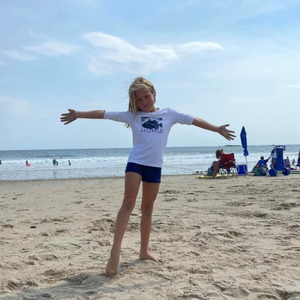Child wearing the white Humu Juniors Classic Long-Sleeve Unisex Rash Guard while standing on the beach