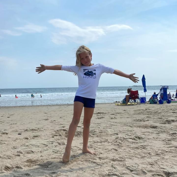 Child wearing the white Humu Juniors Classic Long-Sleeve Unisex Rash Guard while standing on the beach