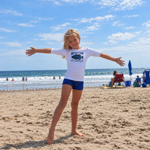 Child wearing the white Humu Juniors Classic Long-Sleeve Unisex Rash Guard while standing on the beach