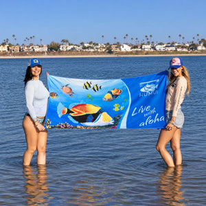 Two women wearing Humu Surf apparel and trucker hats holding the blue Live With Aloha Yoga Towel in shallow ocean water, displaying colorful tropical fish artwork and Humu Surf branding