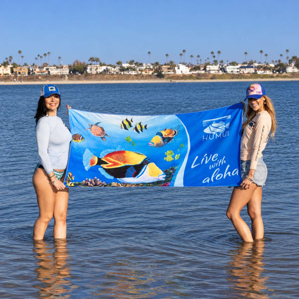 Two women wearing Humu Surf apparel and trucker hats holding the blue Live With Aloha Yoga Towel in shallow ocean water, displaying colorful tropical fish artwork and Humu Surf branding