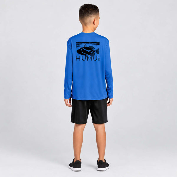 Boy modeling the Maui Ocean Blue Kids Rash Guard from behind, showing the large black Humu Surf fish logo across the back