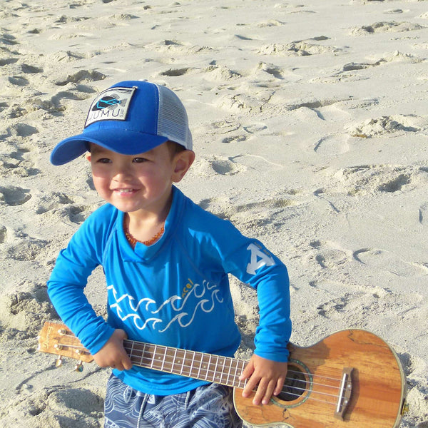 Child wearing the blue Humu Molokini Trucker Hat while holding a ukulele on the beach