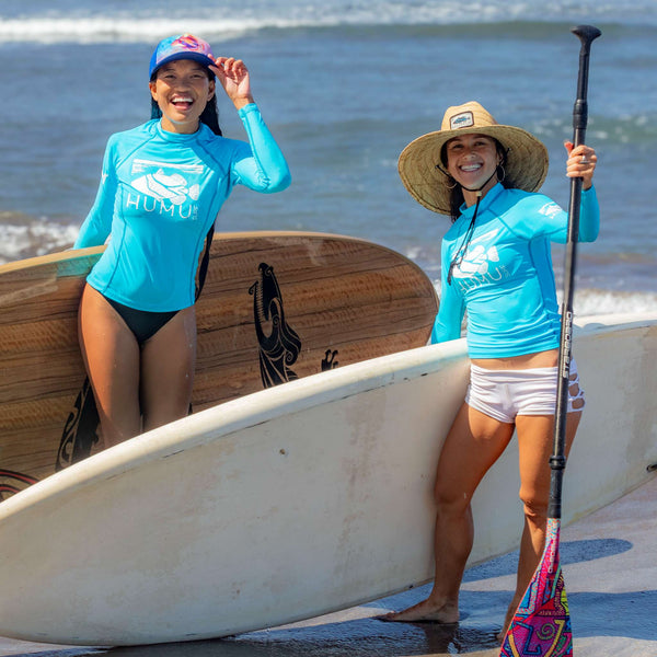 Two women in blue Humu Wahine Kore-Dry Long Sleeve Rashguards with paddleboards, wearing a Napili Trucker Hat and Classic Lifeguard Hat