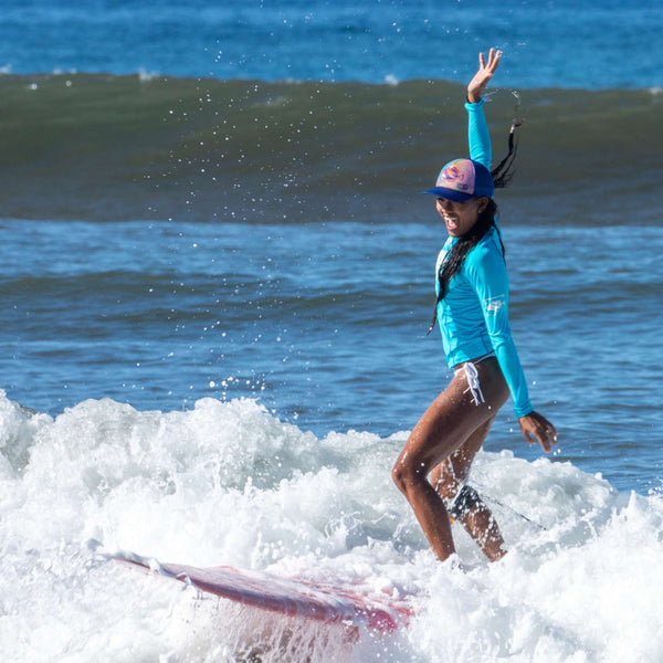 Woman surfing in the blue Humu Wahine Kore-Dry Long Sleeve Rashguard and colorful Napili Trucker Hat