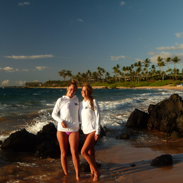 Two women wearing white Humu Women's Woody Long Sleeve Performance Hoodies on a rocky beach beside the ocean