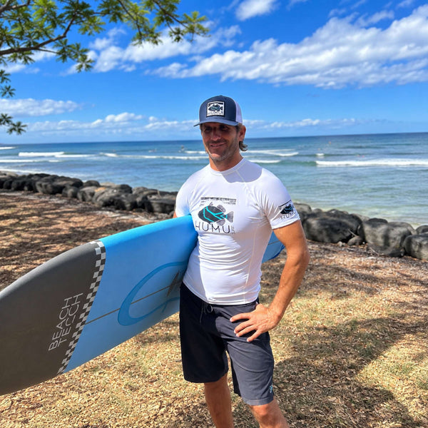 Man wearing a navy and white Humu Woody Trucker Hat and white Humu Surf rash guard while holding a blue surfboard near the ocean