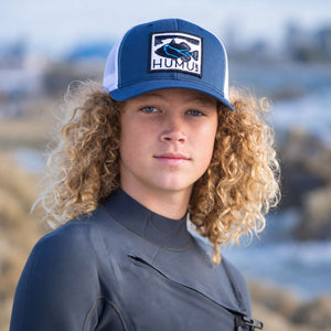 Young surfer wearing the Humu Woody Truckers Hat in navy blue and white with the embroidered Humu Surf fish logo and white mesh back