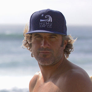 Man wearing the navy and white Humu Woody Trucker Hat beside the ocean with a surfboard