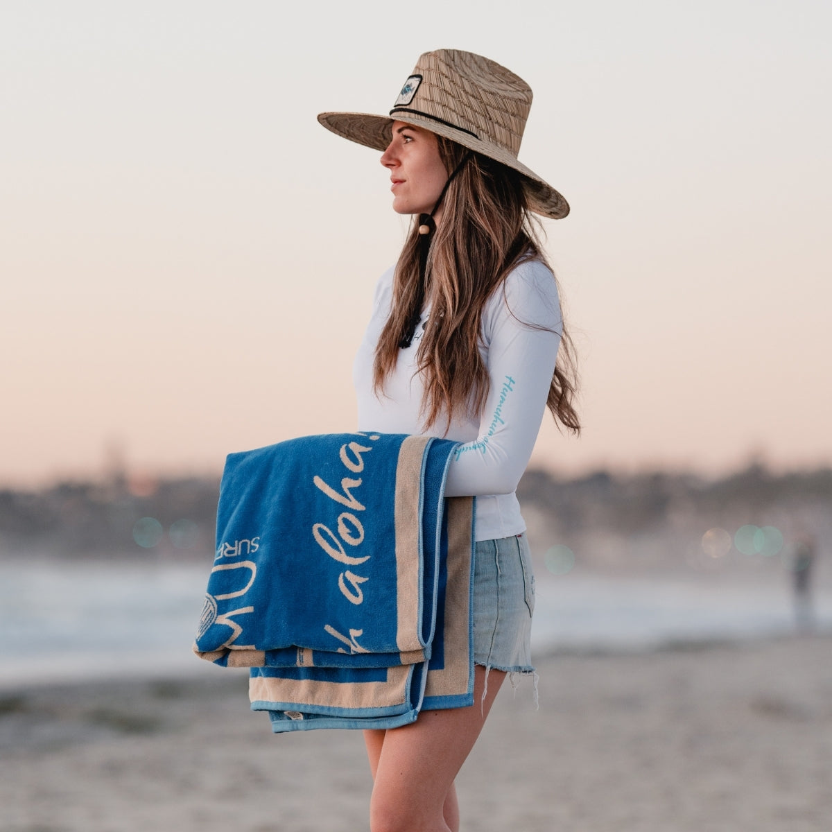 Side profile of a woman wearing the Humu Classic Lifeguard Hat on the beach while carrying a folded blue Humu Surf brand Live With AlohaSurf towel