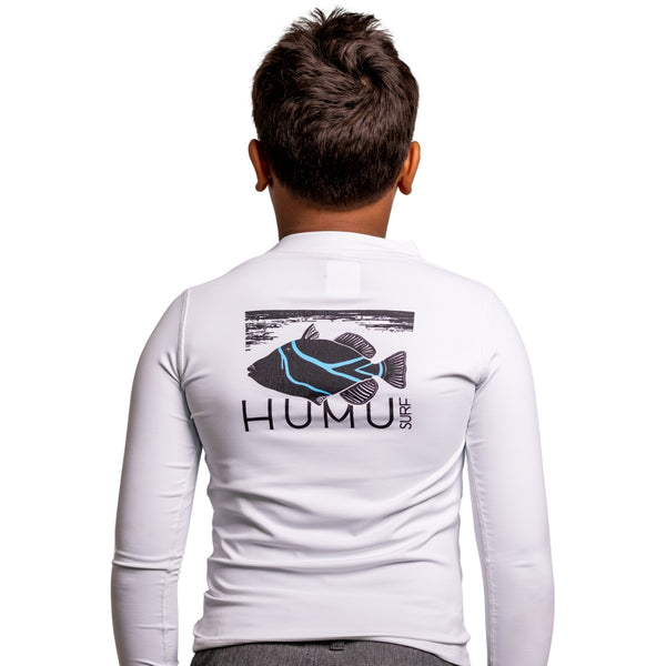 Back view of a boy wearing the white Humu Juniors Classic Long Sleeve Rash Guard showing the large black and blue Humu Surf fish graphic