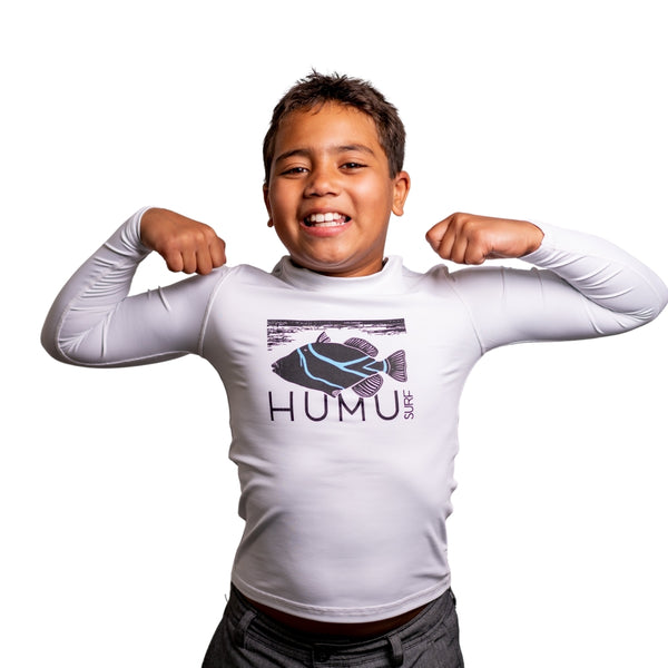 Boy flexing his arms while wearing the white Humu Juniors Classic Long Sleeve Rash Guard with black and blue Humu Surf fish graphic