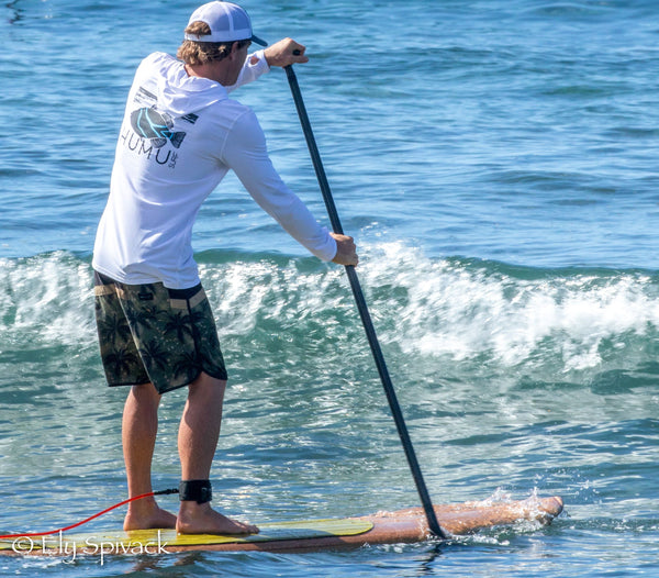 Man wearing the white Humu Men's Woody Long Sleeve Performance Hoody while stand-up paddleboarding on an ocean wave, showing the large Humu fish graphic on the back