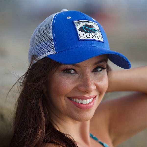 Woman wearing the blue Humu Molokini Trucker Hat with white mesh back and embroidered fish patch