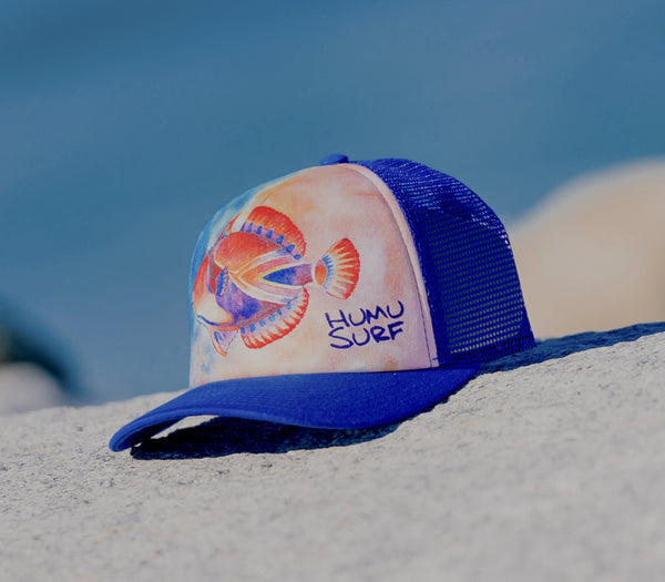 Humu Napili Trucker Hat with colorful fish artwork photographed beside the ocean